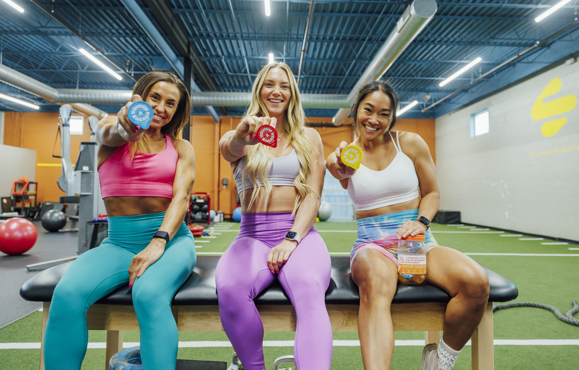 Three fit women in a gym holding energy-boosting pre-workout shots by Squish Energy.