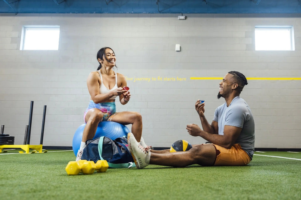 Two people in a gym setting, one on an exercise ball, interacting with sports balls.