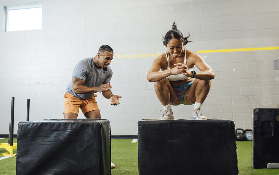 A woman performing a box jump as a fitness trainer motivates her.